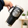 Una mano sostiene un Termo personalizado clásico grande con agarradera 890ml 30oz, una taza de viaje negra con un gráfico de camión y nombre