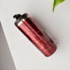 Un termo cilindro metalizado mediano personalizado de 450 ml (15 oz) con cuerpo rojo y tapa negra descansa sobre una superficie blanca junto a una hoja verde.