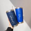 En una mano sostiene dos vasos Termo personalizado clásico de 590ml y 20oz, personalizados con “SARAHÍ” y “Maestro ÁNGEL LÓPEZ”, regalos únicos para cualquier ocasión.