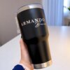 Una mano sostiene un Termo personalizado clásico grande 890ml 30oz, un vaso negro con "ARMANDO Bravo" impreso en letras blancas.