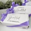 Caja personalizada con 2 diseños kraft mate, decorada con lazo morado y texto en español, se muestra frente a plantas verdes. Ideal para regalos, esta caja ofrece una opción elegante frente a los diseños kraft tradicionales.