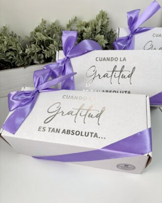 Caja personalizada con 2 diseños kraft mate, decorada con lazo morado y texto en español, se muestra frente a plantas verdes. Ideal para regalos, esta caja ofrece una opción elegante frente a los diseños kraft tradicionales.