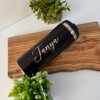 Termo cilindro metalizado mediano 450ml 15oz personalizado en color negro con la palabra "Tanya" escrita en él, expuesto sobre una tabla de madera junto a dos plantas verdes.
