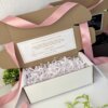 Caja abierta personalizada con 2 diseños kraft mate, decorada con listón rosa, papel relleno y una nota para peticion de padrinos; las plantas de fondo realzan el encanto del diseño.