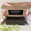 Caja abierta personalizada con 2 diseños kraft mate, rellena con papel negro cortado y decorada con cinta rosa. Un mensaje en español para peticion de padrinos adorna la tapa, mientras una planta verde resalta el estilo de la caja.