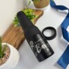 Un portabotellas y lata personalizado de color negro, con cuello y abridor, se encuentra sobre una mesa con plantas, una tabla de madera, una cinta azul y un abridor cerca.