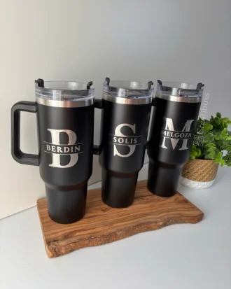 Tres tazas de viaje personalizadas Termo personalizado jumbo con agarradera de 1200 ml (40 oz), cada una de color negro con tapa transparente y etiquetadas con nombres, descansan sobre una bandeja de madera.