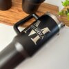 Termo personalizado jumbo con agarradera 1200ml (40oz) de color negro, con una gran letra M y el texto Melgoza, colocado sobre una mesa junto a una planta y una bandeja de madera.
