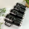 Cuatro vasos negros Termo personalizado clásico grande con agarradera de 890 ml y 30 oz con bordes plateados y nombres blancos se exhiben cerca de plantas verdes.