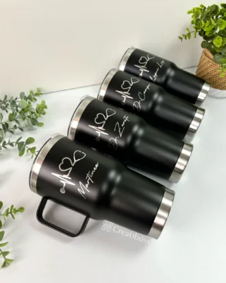 Cuatro vasos negros Termo personalizado clásico grande con agarradera de 890 ml y 30 oz con bordes plateados y nombres blancos se exhiben cerca de plantas verdes.