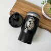 El Termo personalizado portalatas 4 en 1 420 ml 14 oz es un elegante vaso negro con un logotipo y una tapa desmontable, colocado sobre una tabla de madera al lado de una planta, que ofrece un almacenamiento versátil de bebidas.