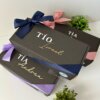 Tres Caja personalizada con 2 diseños kraft mate con nombres y cintas, apiladas junto a pequeñas plantas verdes sobre una superficie blanca.