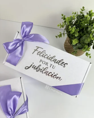 Caja personalizada con 2 diseños kraft mate, decorada con listón morado y la frase "Felicidades por tu jubilación" en la tapa, junto a una maceta.