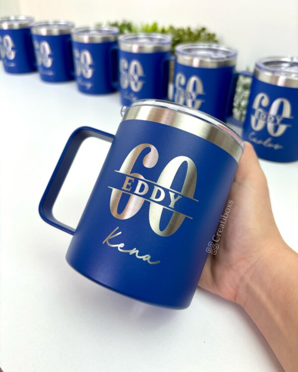 En una mano se sostiene una taza Termo personalizada azul de 360 ml (12 oz) etiquetada como Eddy 60 Kena, con tazas similares en el fondo.