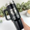 Una mano sostiene un Termo personalizado jumbo con agarradera de 1200 ml (40 oz), una taza de viaje negra con el logo de una cara de oso y el texto "Joes Rail Range". Se ven plantas al fondo.