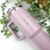 Un Termo personalizado jumbo con agarradera 1200ml 40oz en color rosa se encuentra sobre una superficie blanca, rodeado de hojas verdes.