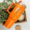 El Termo personalizado jumbo con agarradera 1200ml 40oz en color naranja brillante con logotipo se encuentra junto a una vela y vegetación en una canasta tejida sobre una superficie blanca.