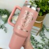 Una mano sostiene un Termo personalizado jumbo con agarradera 1200ml 40oz en color rosa, con un dibujo de un corazón y el nombre Mariana, rodeado de plantas.