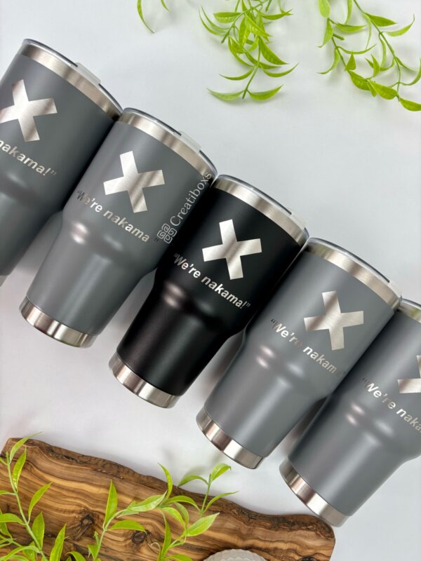 Seis vasos Termo personalizado clásico grande de 890 ml y 30 oz en negro y gris con la inscripción “¡Were nakama!” grabada se exhiben sobre una mesa con hojas verdes, perfectos para disfrutar de tus bebidas favoritas.