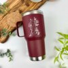Un Termo personalizado clásico grande con agarradera granate de 890 ml y 30 oz con un diseño floral blanco y el texto “Liz”, rodeado de plantas sobre una mesa.