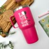 Un Termo personalizado clásico grande con agarradera rosa de 890 ml y 30 oz, con "LOVE Maestra Marcela" en texto decorativo, se muestra sobre una superficie blanca con plantas y cuadernos.