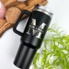 El Termo personalizado jumbo con agarradera 1200ml 40oz, etiquetado M Miranda en color negro, se encuentra sobre un escritorio con plantas, un posavasos y una vela rosa cerca; perfecto para mantener las bebidas calientes o frías.