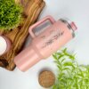Termo personalizado jumbo con agarradera 1200ml 40oz color rosa, con diseño floral, sobre bandeja de madera, rodeado de plantas verdes y una vela.