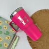 Un termo personalizado clásico grande con agarradera de 890ml, etiquetado como Lorena y en color rosa, está sobre una alfombrilla junto a bolígrafos y un cuaderno con estampado de aguacate.