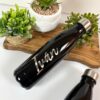Una elegante botella de termo personalizada de 500 ml y 17 oz de color negro con el nombre Iván grabado con elegancia reposa sobre una superficie de madera. Alrededor de la botella personalizada hay plantas en macetas de cerámica tejida y blanca, mientras que otra botella similar se asoma a la vista.