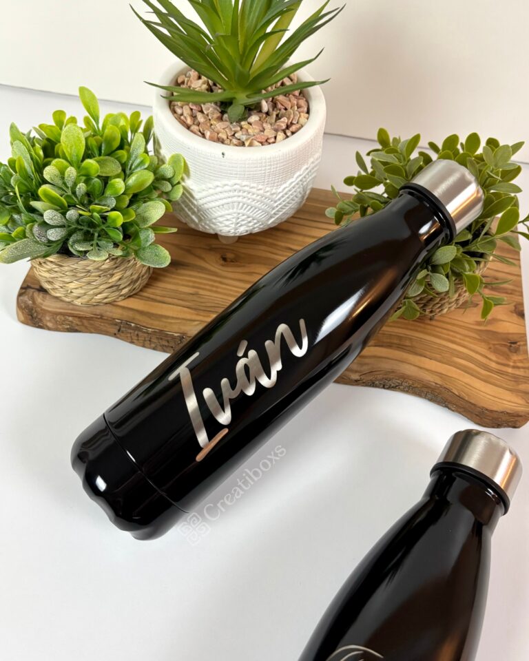 Una elegante botella de termo personalizada de 500 ml y 17 oz de color negro con el nombre Iván grabado con elegancia reposa sobre una superficie de madera. Alrededor de la botella personalizada hay plantas en macetas de cerámica tejida y blanca, mientras que otra botella similar se asoma a la vista.