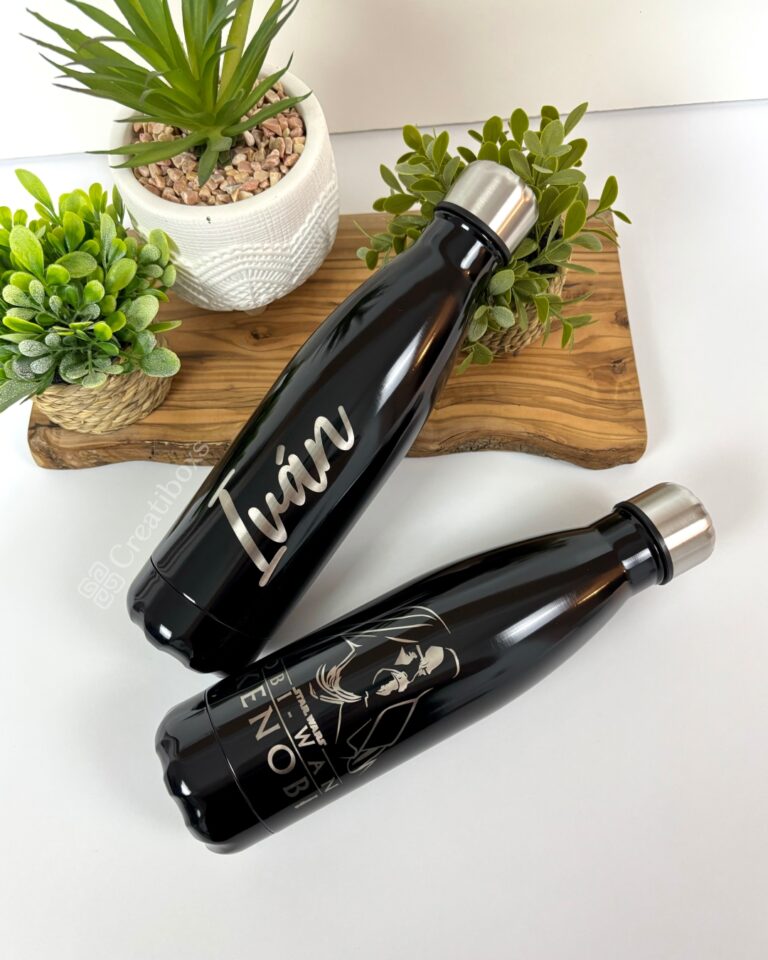 Dos botellas negras de la marca "Termo botella personalizado 500ml" descansan elegantemente sobre una superficie blanca. Una lleva el nombre de Iván en blanco, mientras que la otra muestra un personaje con el texto KENOBI. Ambas están colocadas elegantemente junto a pequeñas plantas verdes.