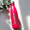Una botella Termo personalizada de 500 ml (17 oz) de color rosa, con la palabra "Barbie" en negrita, reposa sobre una superficie blanca parcialmente en sombra. Cerca hay una ramita de hojas verdes. Este accesorio combina a la perfección estilo y funcionalidad, añadiendo un toque de encanto a cualquier entorno.