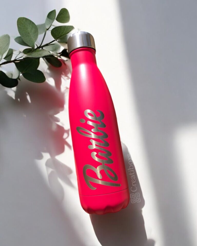 Una botella Termo personalizada de 500 ml (17 oz) de color rosa, con la palabra "Barbie" en negrita, reposa sobre una superficie blanca parcialmente en sombra. Cerca hay una ramita de hojas verdes. Este accesorio combina a la perfección estilo y funcionalidad, añadiendo un toque de encanto a cualquier entorno.