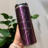 Una mano sostiene un termo cilindro metalizado mediano de 450 ml personalizado de color morado con un diseño estilizado de la firma. En el fondo, una planta frondosa añade un toque de naturaleza, mientras que la persona que lleva una pulsera beige muestra su vaso con estilo.