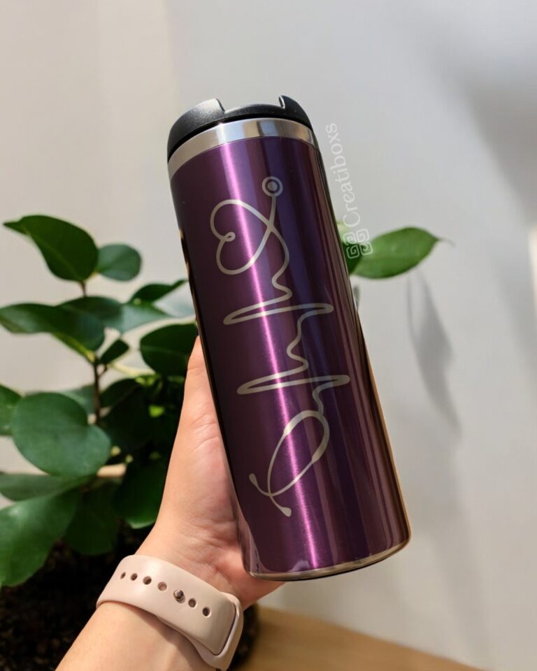 Una mano sostiene un termo cilindro metalizado mediano de 450 ml personalizado de color morado con un diseño estilizado de la firma. En el fondo, una planta frondosa añade un toque de naturaleza, mientras que la persona que lleva una pulsera beige muestra su vaso con estilo.