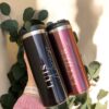 Dos termos de acero inoxidable personalizados, etiquetados con los nombres de Luis y Esther, se sostienen sobre un fondo verde exuberante de hojas de eucalipto. El "Termo cilindro metalizado mediano personalizado 450ml 15oz" de la izquierda es negro, mientras que el de la derecha es rosa metalizado.