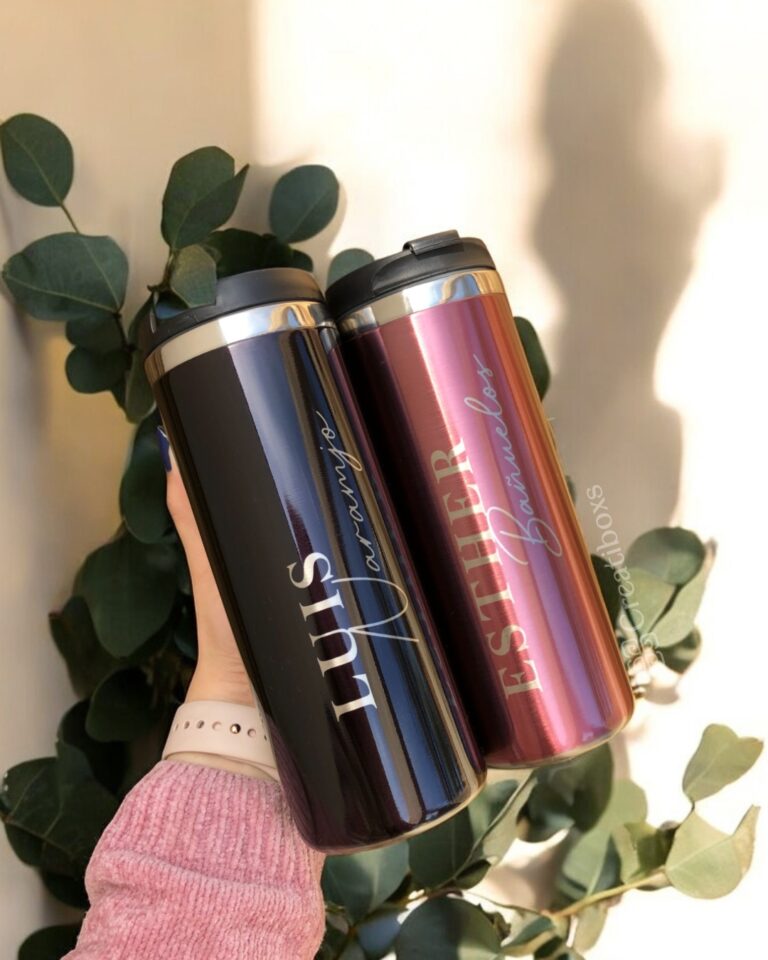 Dos termos de acero inoxidable personalizados, etiquetados con los nombres de Luis y Esther, se sostienen sobre un fondo verde exuberante de hojas de eucalipto. El "Termo cilindro metalizado mediano personalizado 450ml 15oz" de la izquierda es negro, mientras que el de la derecha es rosa metalizado.