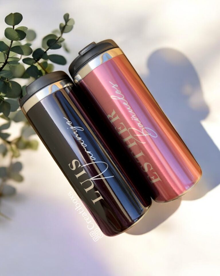 Dos vasos de viaje medianos personalizados de metal, cada uno con tapa, se encuentran en posición vertical. Uno de los vasos presenta un acabado oscuro con la palabra "LUIS" elegantemente escrita en blanco, mientras que el otro brilla en oro rosa y muestra la palabra "ESTHER" en una escritura a juego. Una ramita de eucalipto reposa junto a estos elegantes "Termo cilindro metalizado mediano personalizado 450ml 15oz" sobre un fondo claro.