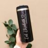 Una mano sostiene un elegante termo cilindro metalizado mediano personalizado de 450 ml y 15 oz de color negro, con la inscripción "Eris Elizabeth Geduraryrama" en cursiva blanca. El vaso personalizado se encuentra sobre un fondo beige adornado con hojas de eucalipto verde a la izquierda. La persona lleva un elegante reloj inteligente blanco.