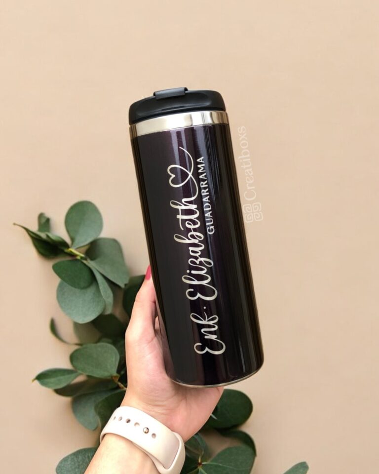 Una mano sostiene un elegante termo cilindro metalizado mediano personalizado de 450 ml y 15 oz de color negro, con la inscripción "Eris Elizabeth Geduraryrama" en cursiva blanca. El vaso personalizado se encuentra sobre un fondo beige adornado con hojas de eucalipto verde a la izquierda. La persona lleva un elegante reloj inteligente blanco.