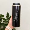 Una mano sostiene un elegante termo cilindro metalizado mediano personalizado en color negro, rematado con una tapa plateada pulida. La taza de viaje muestra los nombres Vizquet y Sergio con elegancia en letras blancas cursivas y mayúsculas. Una planta verde frondosa proporciona un hermoso fondo, mientras que la marca de agua de Creatboxes la destaca discretamente como una verdadera obra maestra.