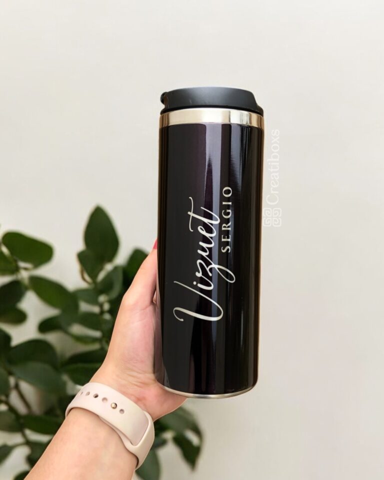 Una mano sostiene un elegante termo cilindro metalizado mediano personalizado en color negro, rematado con una tapa plateada pulida. La taza de viaje muestra los nombres Vizquet y Sergio con elegancia en letras blancas cursivas y mayúsculas. Una planta verde frondosa proporciona un hermoso fondo, mientras que la marca de agua de Creatboxes la destaca discretamente como una verdadera obra maestra.