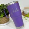 Un Termo clásico grande con agarradera personalizada de 890 ml (30 oz) en color morado, adornado con el nombre "Julia" elegantemente escrito en plata, adorna la escena. Con una base clásica de plata y un asa personalizada, se encuentra sobre una superficie blanca junto a una canasta tejida, una pequeña planta y una vela perfumada que brilla suavemente.