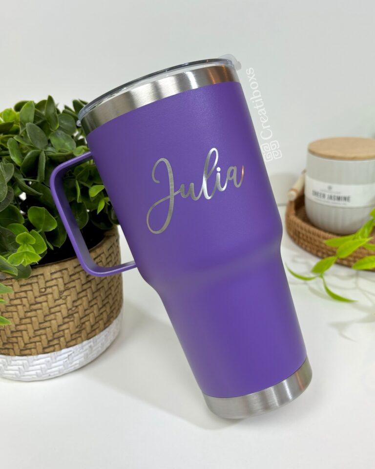 Un Termo clásico grande con agarradera personalizada de 890 ml (30 oz) en color morado, adornado con el nombre "Julia" elegantemente escrito en plata, adorna la escena. Con una base clásica de plata y un asa personalizada, se encuentra sobre una superficie blanca junto a una canasta tejida, una pequeña planta y una vela perfumada que brilla suavemente.