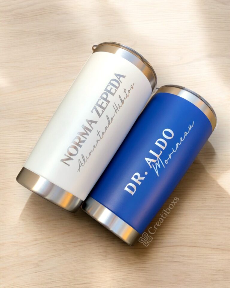 Dos vasos Termo clásico personalizado de 590 ml se encuentran sobre una superficie de madera. El vaso blanco muestra "Norma Zepeda, Alimentando Hábitos", mientras que el azul muestra "Dr. Aldo Mondaca". Ambos tienen tapas y bases de metal, lo que ejemplifica el diseño clásico personalizado del termo.