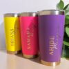 Se muestran tres vasos personalizados Termo clásico en colores vibrantes: uno amarillo con el nombre BRIANA, otro rosa con el nombre SAMARA y uno morado con la etiqueta AHIDE. Cada vaso incluye una base y un borde superior metálicos, con un fondo de plantas frondosas.