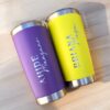 Dos vasos de acero inoxidable, con exteriores vibrantes, descansan sobre una superficie de madera. El vaso morado de la izquierda muestra "AHIDE" y "Mangana" en letras blancas, mientras que el vaso amarillo de la derecha muestra "BRIANA" y "Mega" en azul. Ambos son ejemplos del Termo clásico personalizado de 590 ml (20 oz).
