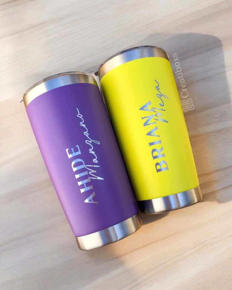 Dos vasos de acero inoxidable, con exteriores vibrantes, descansan sobre una superficie de madera. El vaso morado de la izquierda muestra "AHIDE" y "Mangana" en letras blancas, mientras que el vaso amarillo de la derecha muestra "BRIANA" y "Mega" en azul. Ambos son ejemplos del Termo clásico personalizado de 590 ml (20 oz).