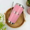 Termo personalizado clásico 590ml 20oz en color rosa, con el nombre Natalia en blanco, sobre una bandeja de mimbre y rodeado de plantas verdes.
