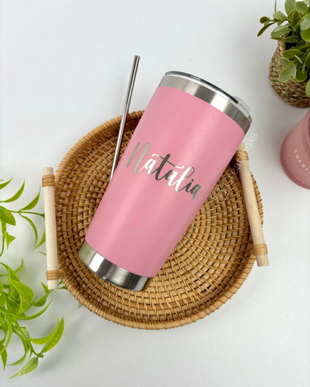 Termo personalizado clásico 590ml 20oz en color rosa, con el nombre Natalia en blanco, sobre una bandeja de mimbre y rodeado de plantas verdes.
